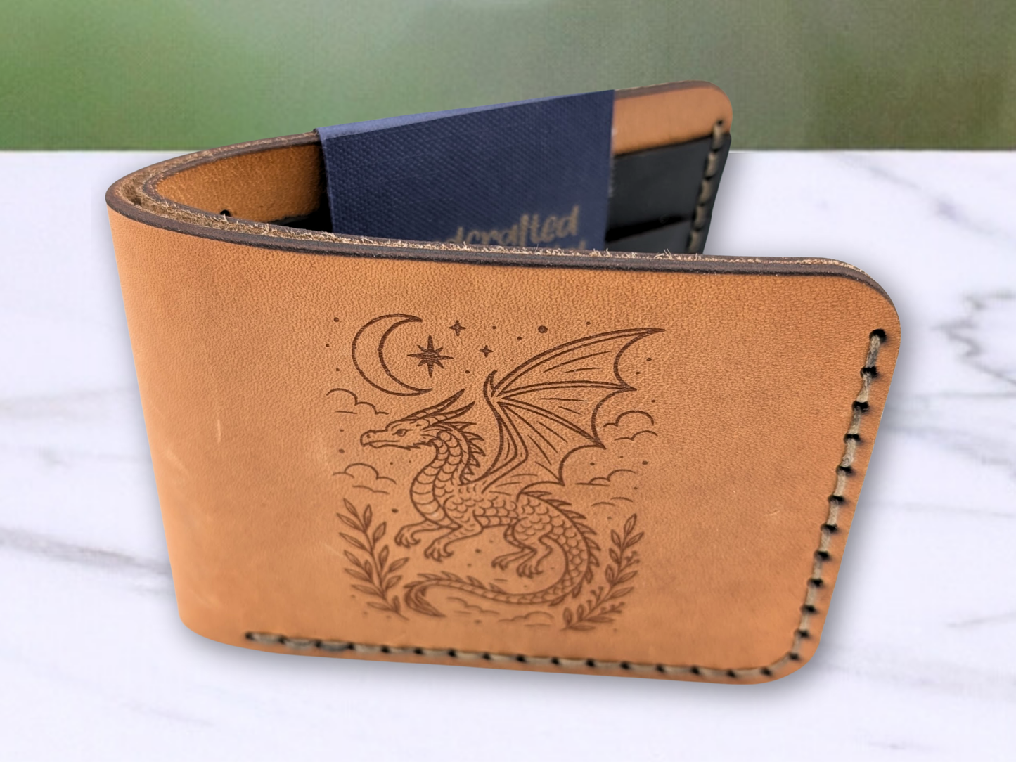 Front view of handcrafted veg tan leather (light brown and not dyed) bi fold wallet. In this photo you are looking at the front of the wallet which is standing on it's side from above. You can see the leather has darkened edges, the two layers of leather that form the cash compartment, stitching, laser engraved dragon, and black card pockets can be glimpsed in the interior. The dragon has wings extended and is surrounded by clouds with a crescent moon and star above the dragon. 