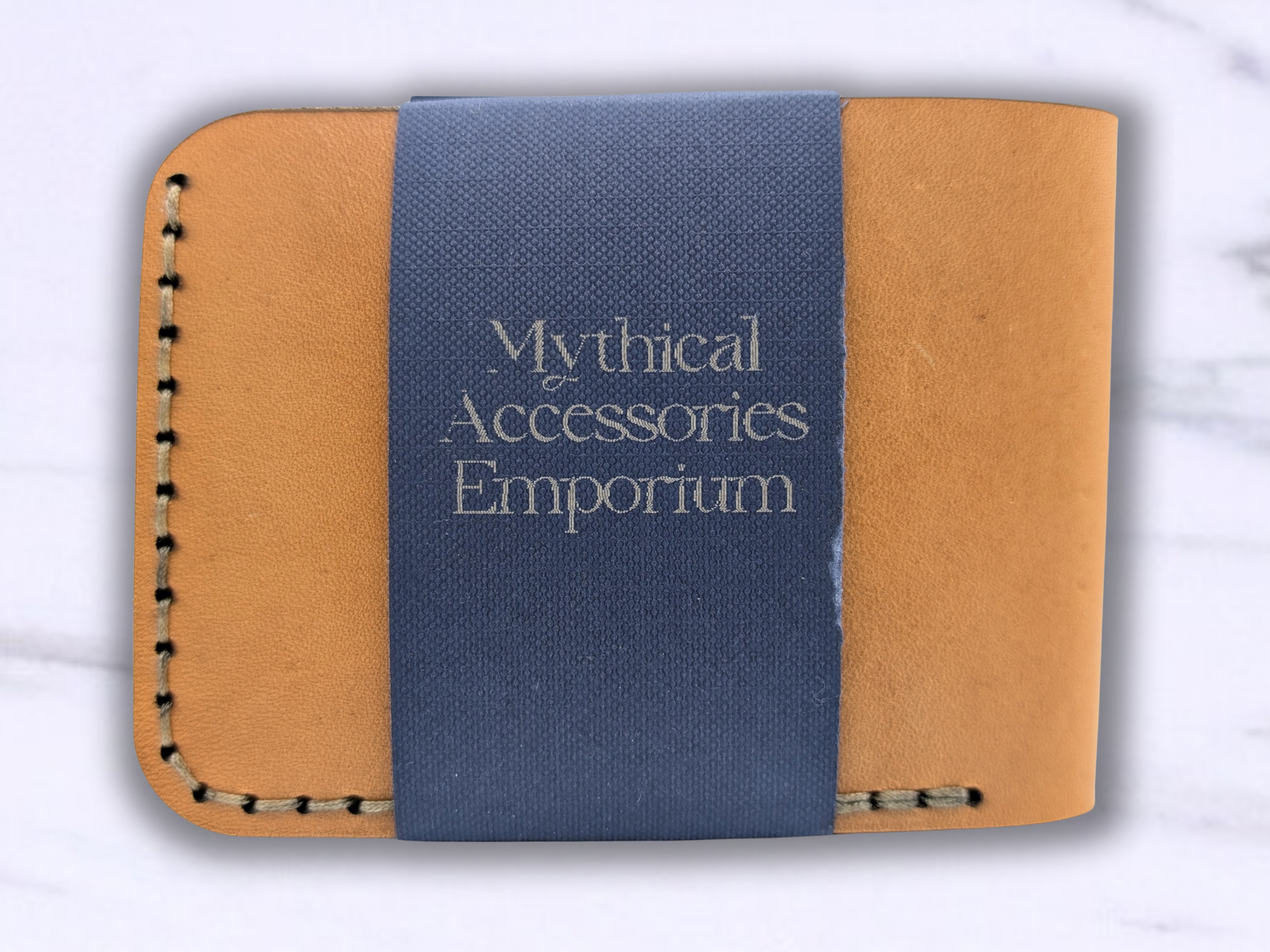 View of the dragon leather bi fold wallet. Here you are looking at the back of the wallet which is a smooth tan surface with stitching. The Mythical Accessories Emporium packaging in a dark blue stripe wraps around the back.