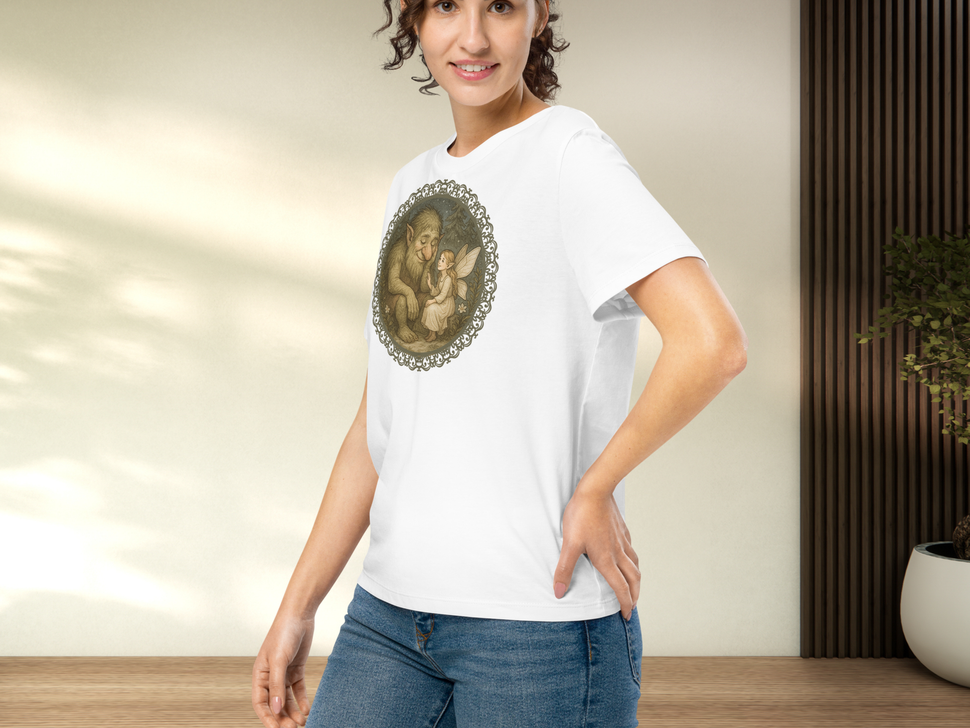 Model wearing a white graphic fairy t-shirt.  The model's hand is on her hip and you can see the side and front of the shirt. This pose allows you to see the side seam, The fit of the short sleeve and where the hem falls mid hip. The graphic's frame is  lattice lace. Inside the frame is a fairy/pixie sitting in the forest with a forest troll.