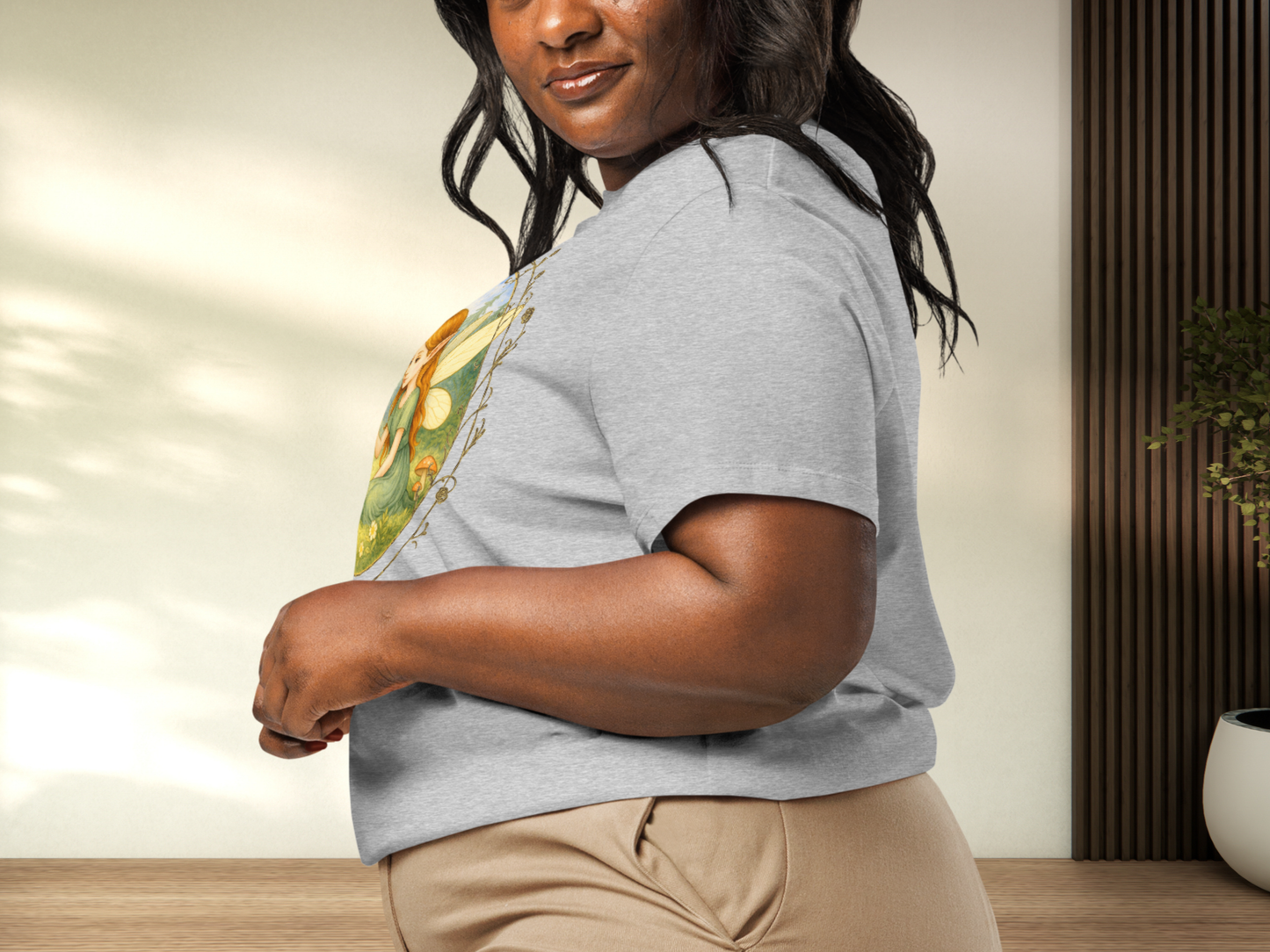 Curvy model wearing an athletic grey t-shirt with a fantasy graphic, left side view. Circle shaped graphic is framed in gold rose vines. A fairy kneels in a forest meadow petting a reddish baby dragon. Fairy is wearing a long green dress and has auburn hair. Insect like wings. 