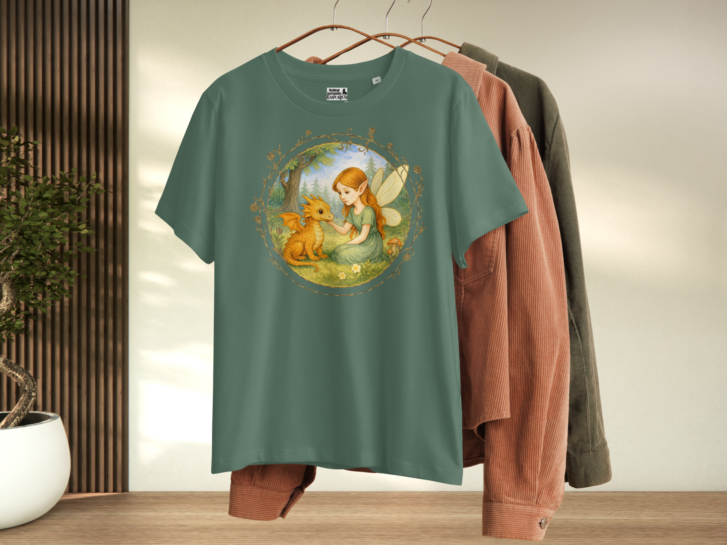 Bay Green t-shirt with a fantasy graphic hanging with other shirts. Circle shaped graphic is framed in gold rose vines. A fairy kneels in a forest meadow petting a reddish baby dragon. Fairy is wearing a long green dress and has auburn hair. Insect like wings. 