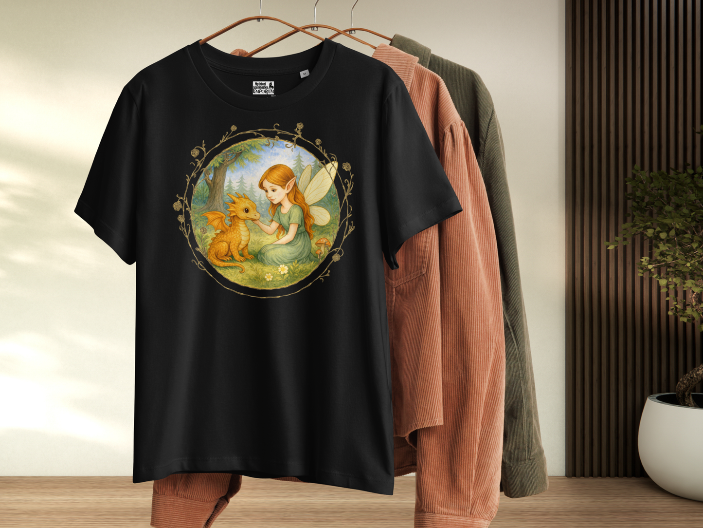 Black women's fantasy tee hanging with other shirts. Circle shaped graphic is framed in gold rose vines. A fairy kneels in a forest meadow petting a reddish baby dragon. Fairy is wearing a long green dress and has auburn hair. Insect like wings. 