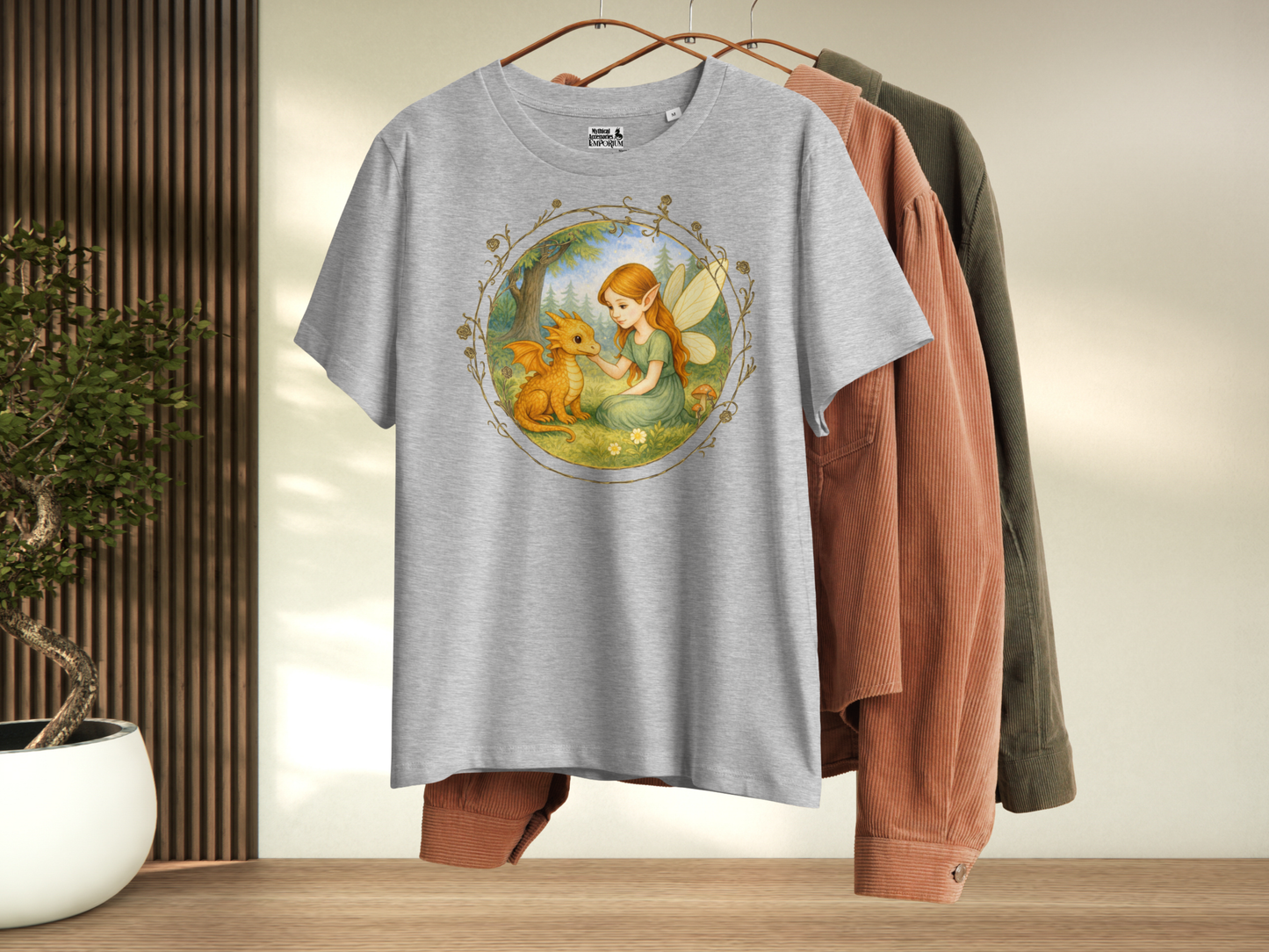 Athletic Grey t-shirt with a fantasy graphic hanging with other shirts. Circle shaped graphic is framed in gold rose vines. A fairy kneels in a forest meadow petting a reddish baby dragon. Fairy is wearing a long green dress and has auburn hair. Insect like wings. 