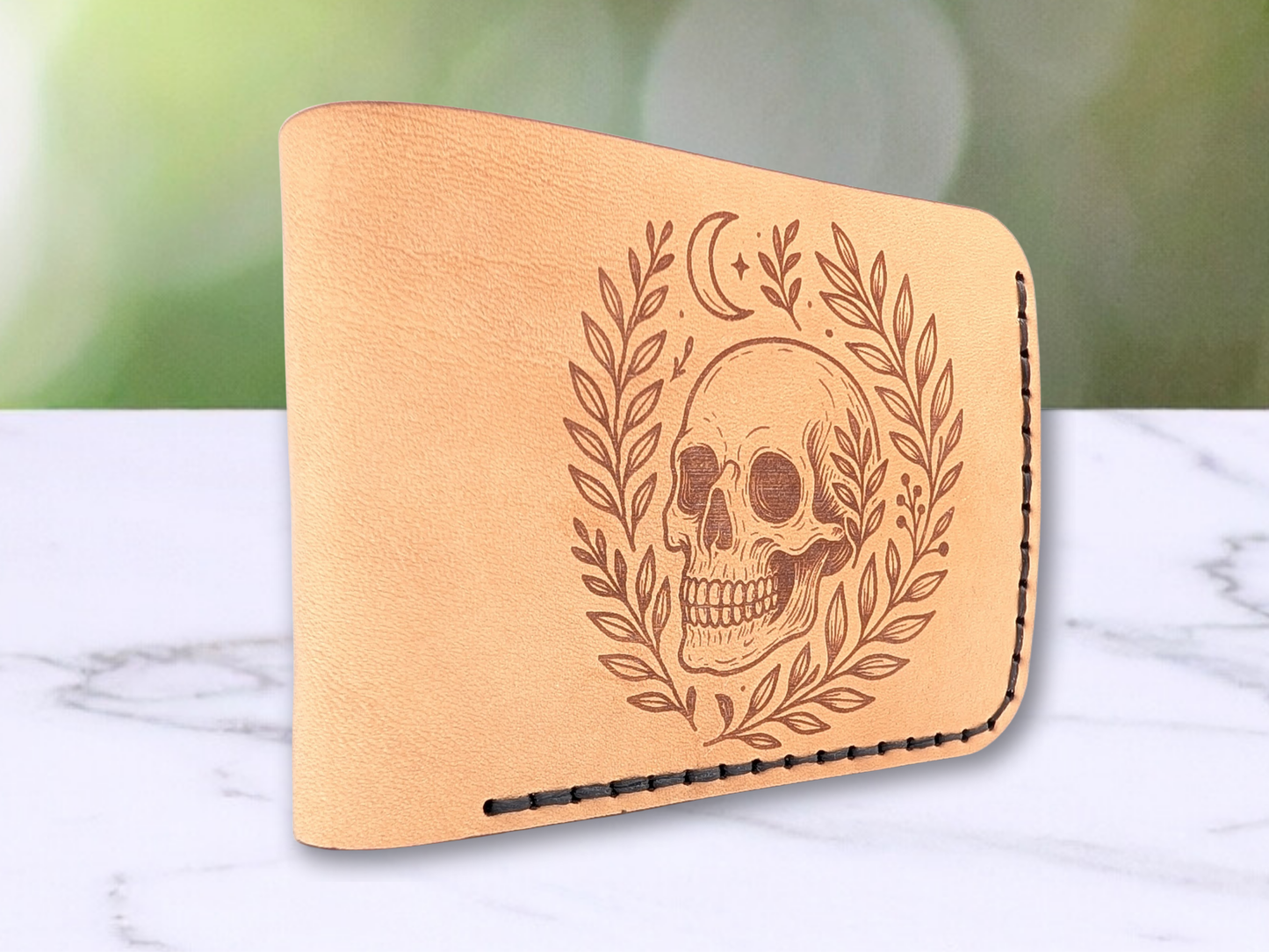 Handmade leather bifold wallet with laser engraved skull. Wallet is standing up with the folded edge closest to the viewer. You see  the rounded edge of the fold.  Front view of wallet with dark stitching on the right side and bottom. Leather is a light brown made from veg tan leather. Detailed skull with a crescent moon above it and vines surrounding both sides of the skull.u