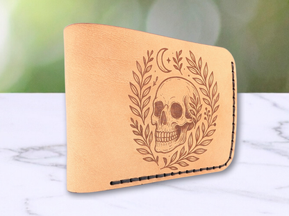 Handmade leather bifold wallet with laser engraved skull. Wallet is standing up with the folded edge closest to the viewer. You see  the rounded edge of the fold.  Front view of wallet with dark stitching on the right side and bottom. Leather is a light brown made from veg tan leather. Detailed skull with a crescent moon above it and vines surrounding both sides of the skull.u