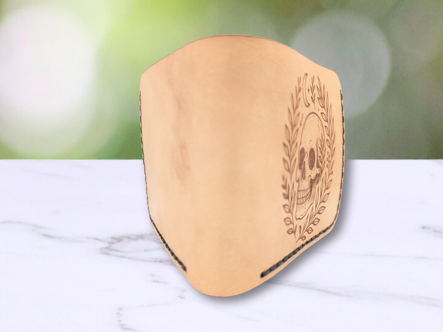 Veg tan handcrafted leather wallet with laser engraved skull standing up with the folded edge facing the view. You glimpse the front and back of the wallet. You see natural color variations in the light brown veg tan leather wallet.