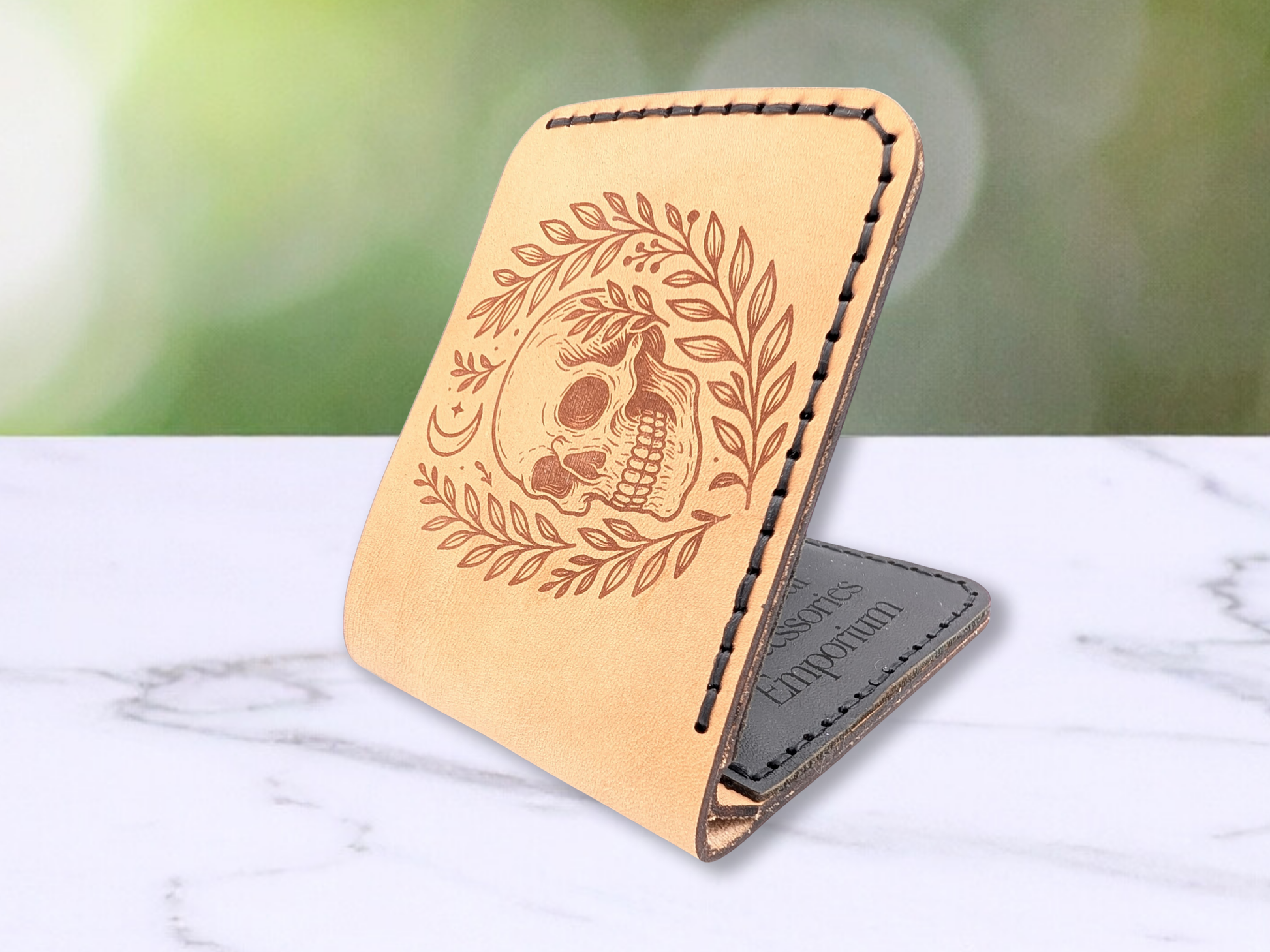 Skull handcrafted leather bifold wallet. Wallet is laying flat on a white marble surface. Wallet is opened so that the front is sticking up. You can see into the interior to see on the black leather card pockets the logo for Mythical Accessories Emporium is engraved. Wallet has black stitching. 