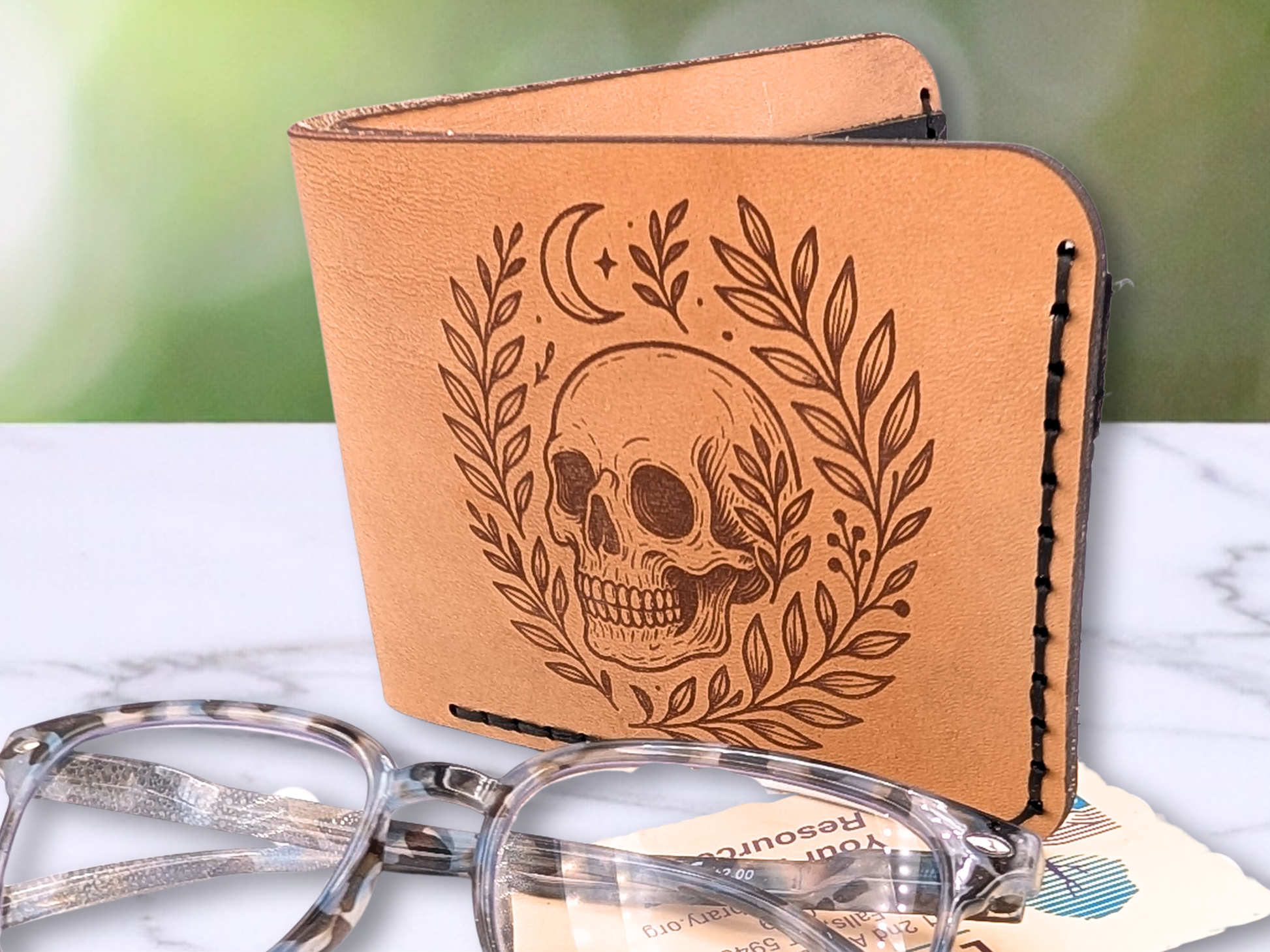 Handmade leather bifold wallet with laser engraved skull. Wallet is standing up slightly opened. Front view of wallet with dark stitching on the right side and bottom. Leather is a light brown made from veg tan leather. Detailed skull with a crescent moon above it and vines surrounding both sides of the skull.