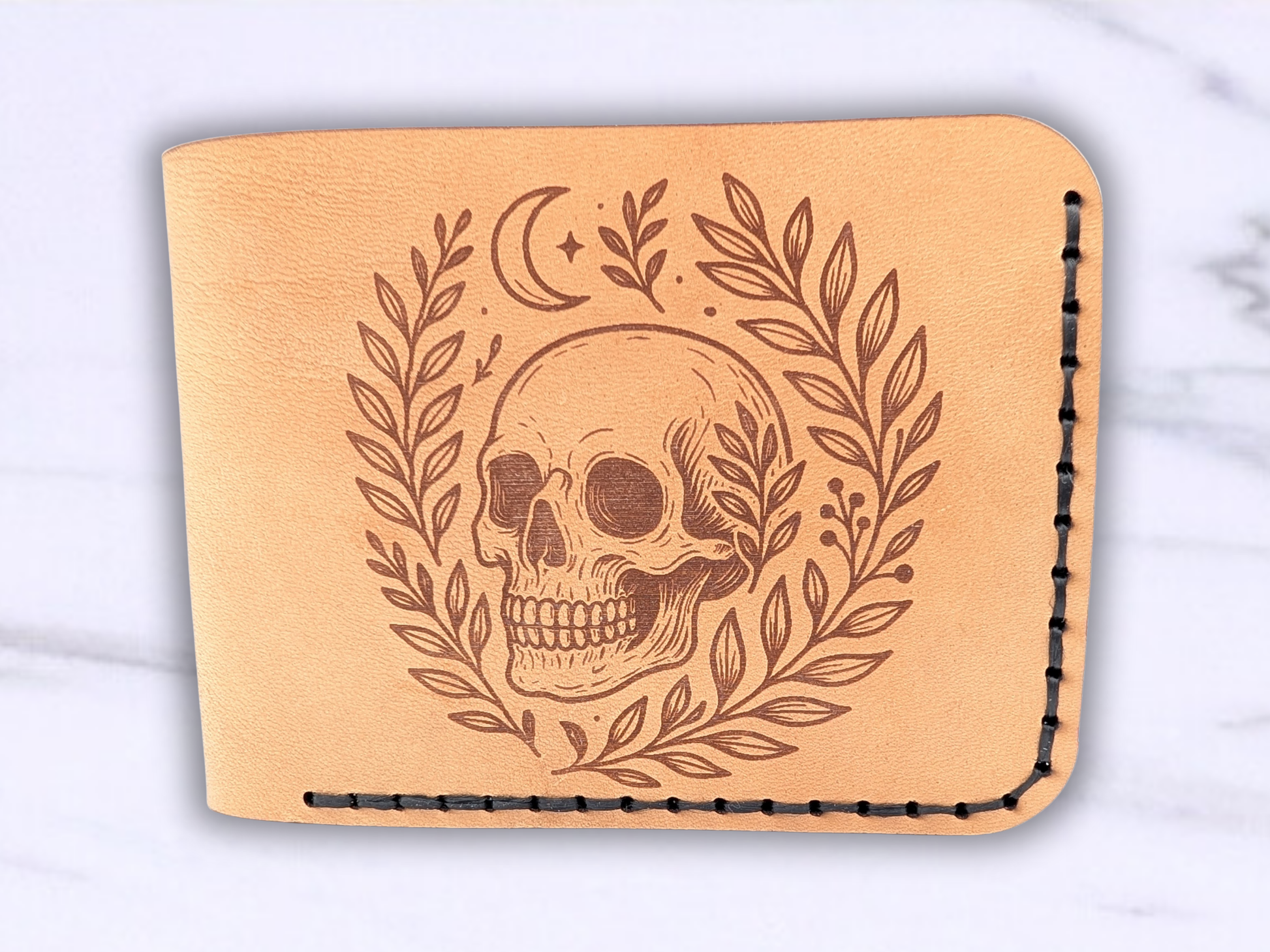Front view of handmade leather bifold wallet with laser engraved skull. Front view of wallet with dark stitching on the right side and bottom. Leather is a light brown made from veg tan leather. Detailed skull with a crescent moon above it and vines surrounding both sides of the skull.