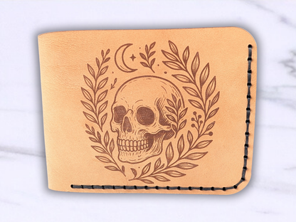 Front view of handmade leather bifold wallet with laser engraved skull. Front view of wallet with dark stitching on the right side and bottom. Leather is a light brown made from veg tan leather. Detailed skull with a crescent moon above it and vines surrounding both sides of the skull.