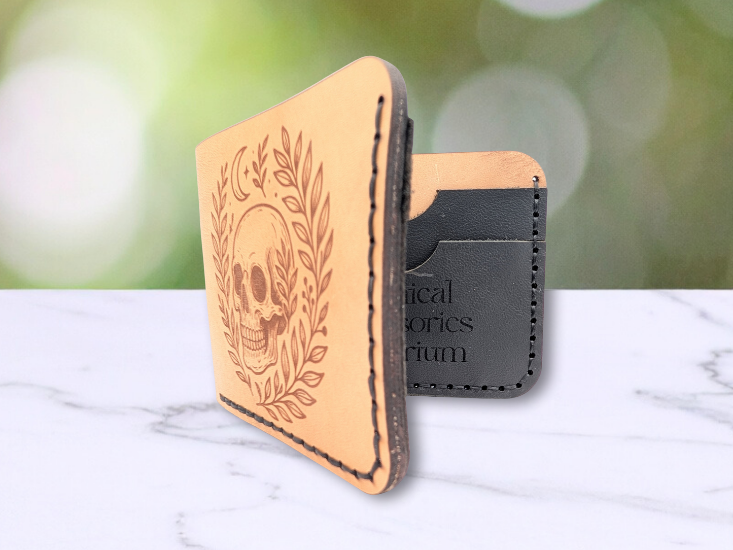 Handmade leather bifold wallet with laser engraved skull. Wallet is standing up opened. Front view of wallet, you can see slightly into the interior to see the black pebbled leather card pockets with black stitching. You can see the darkened edges of the multiple layers of leather that are hand sewn together. Leather is a light brown made from veg tan leather. Detailed skull with a crescent moon above it and vines surrounding both sides of the skull.