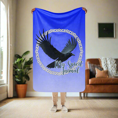 My Spirit Animal Raven Crow Sherpa Blanket. Front View of blanket, 37"x57", held up by model, this is a travel size or a children's size blanket. Raven wings spread as he soars inside a white Celtic Knot circle frame, My Spirit Animal bisects the lower right side of the circle.  Background of blanket is a dip dyed deep violet blue. Darker at the top  with a graduated color to the lighter bottom. Mythical Accessories Emporium. the crow shirt, Morrigan, Celtic Mythology, Odin's Ravens, WIccan