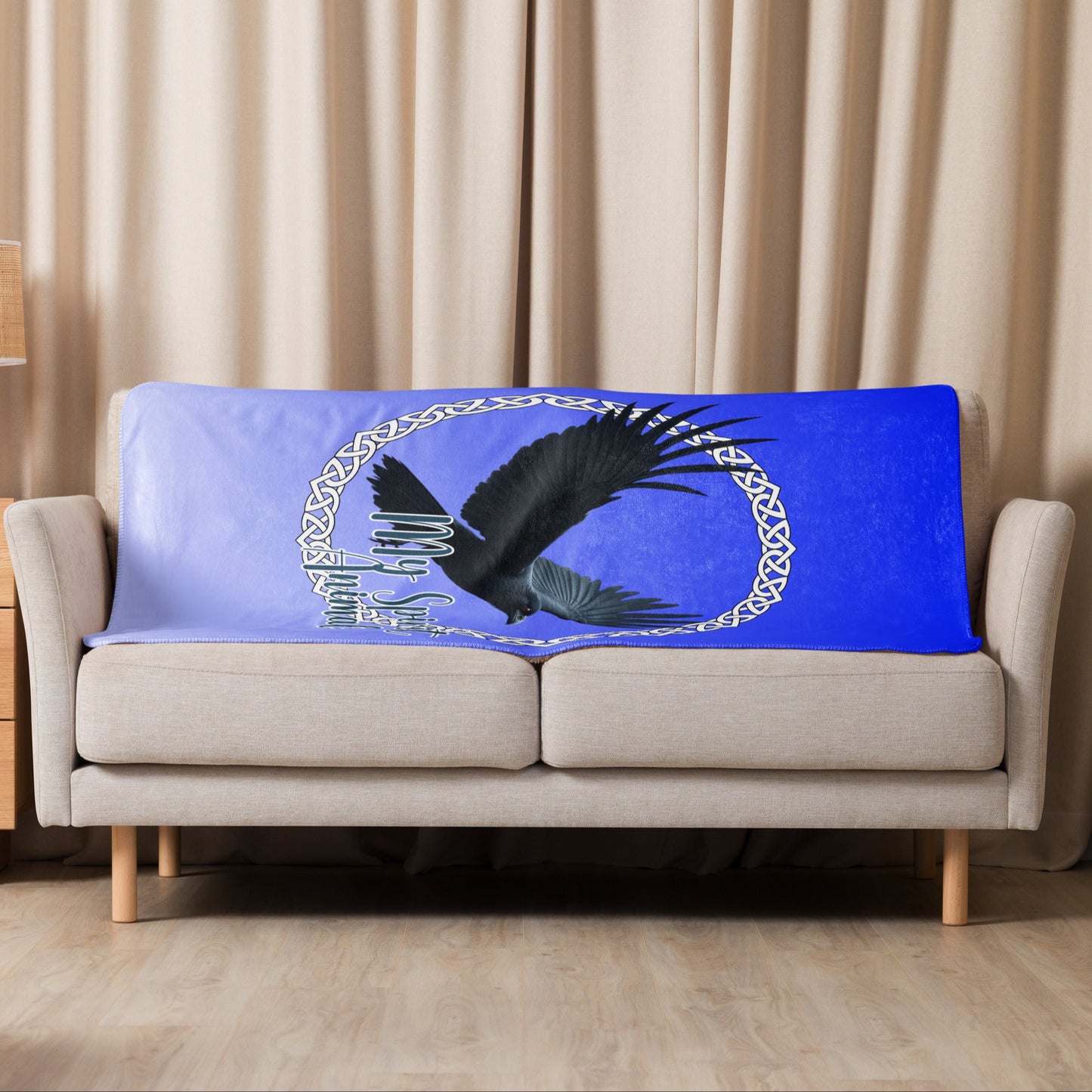 My Spirit Animal Raven Crow Sherpa Blanket. Front View of blanket shown over the back of a loveseat, size 37"x57". Raven wings spread as he soars inside a white Celtic Knot circle frame, My Spirit Animal bisects the lower right side of the circle.  Background of blanket is a dip dyed deep violet blue. Darker at the top  with a graduated color to the lighter bottom. Mythical Accessories Emporium. the crow shirt, Morrigan, Celtic Mythology, Odin's Ravens, WIccan