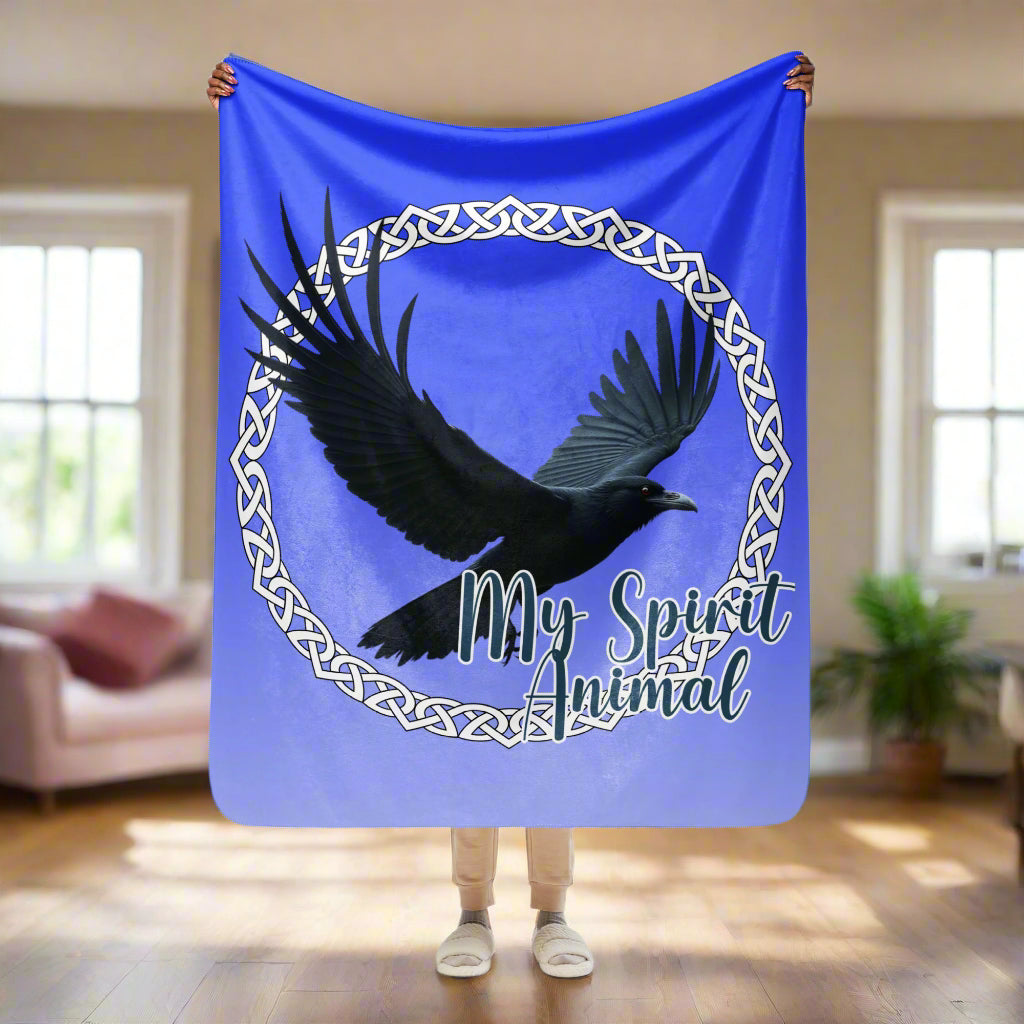 My Spirit Animal Raven Crow Sherpa Blanket. Front View of blanket shown held  up by model, 50x60, bottom is past knees.  Raven wings spread as he soars inside a white Celtic Knot circle frame, My Spirit Animal bisects the lower right side of the circle.  Background of blanket is a dip dyed deep violet blue. Darker at the top  with a graduated color to the lighter bottom. Mythical Accessories Emporium. the crow shirt, Morrigan, Celtic Mythology, Odin's Ravens, WIccan