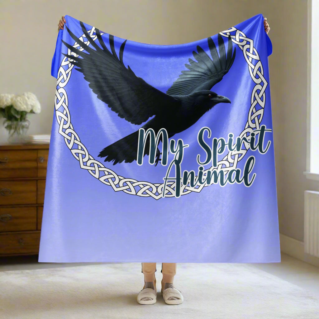 My Spirit Animal Raven Crow Sherpa Blanket. Front View of blanket held up by a model over her heat, 1/4 of blanket is draped over her hands 60x80 twin blanket. Raven wings spread as he soars inside a white Celtic Knot circle frame, My Spirit Animal bisects the lower right side of the circle.  Background of blanket is a dip dyed deep violet blue. Darker at the top  with a graduated color to the lighter bottom. Mythical Accessories Emporium. the crow shirt, Morrigan, Celtic Mythology, Odin's Ravens, WIccan