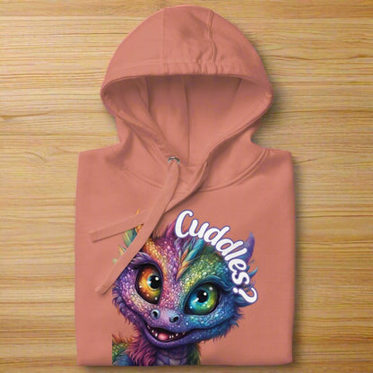 dusty rose Women's Hoodie Front View folded on a wooden surface with hood up. Purple cross eyed baby dragon. Text above dragon circling head "Cuddles?". 