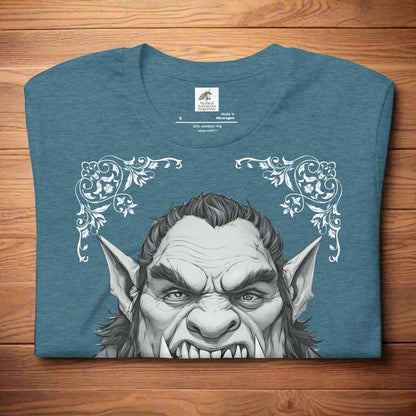 Men's heather deep teal Orc T-Shirt Front View folded on wooden surface. Orc with tusks and pointed ears. Troll TShirt, Giant TShirt, Ogre Shirt