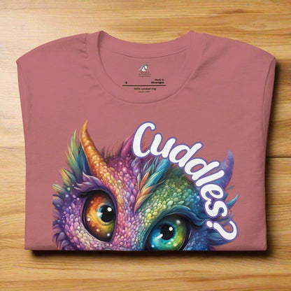 Mauve Dragon Cuddles Front View Men's T-Shirt folded on a wooden surface. Whimsical purple iridescent  dragon looking up at the word Cuddles? above his headockup