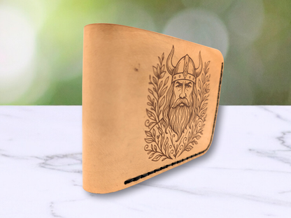 Viking Leather  Handmade Wallet . Front view of wallet with the focus on the folded edge of the wallet. Laser engraved with a stern face of a Viking male warrior with a horned helmet, long beard and hair. He is surrounded with botanical vines on both sides. The laser engraving is a dark brown to black in color. The hand sewn stitching in dark waxed cord can be seen on the right and bottom of the wallet. 