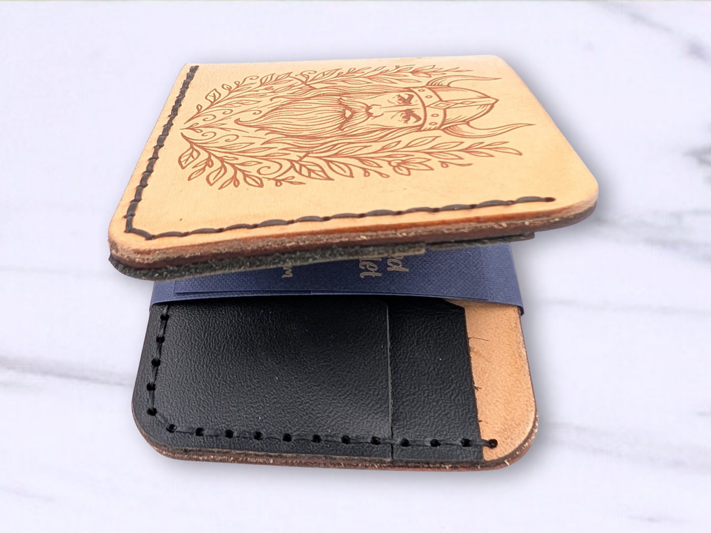 Viking laser engraved handmade bi fold wallet. Wallet is laying flat on a white surface turned so that the side that opens if facing you. You can see the double thickness of the veg tan leather that makes up the main body. Attached to this on the interior you can see the double thickness of the black pebbled leather that makes up the card pockets on each side. Wallet is hand sewn with dark cording which is visible. 