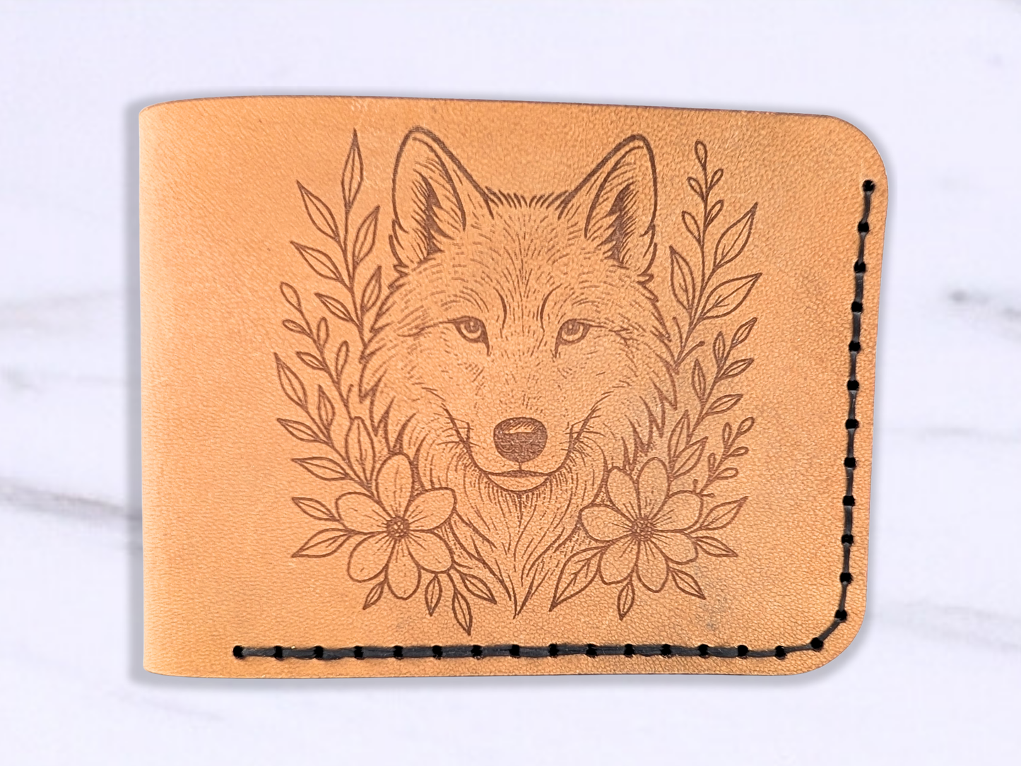 Leather veg tan wallet with laser engraved wolf head looking out at you on the front. To each side of the wolf are vines and flowers. In the photo the wallet is laying flat to see only the front of the light brown leather. The wolf head is looking straight out with ears raised and alert. The wolf is surrounded by vines and flowers. Dark stitching around the bottom and right side of the wallet is visible. 