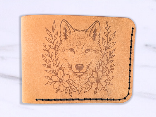 Leather veg tan wallet with laser engraved wolf head looking out at you on the front. To each side of the wolf are vines and flowers. In the photo the wallet is laying flat to see only the front of the light brown leather. The wolf head is looking straight out with ears raised and alert. The wolf is surrounded by vines and flowers. Dark stitching around the bottom and right side of the wallet is visible. 