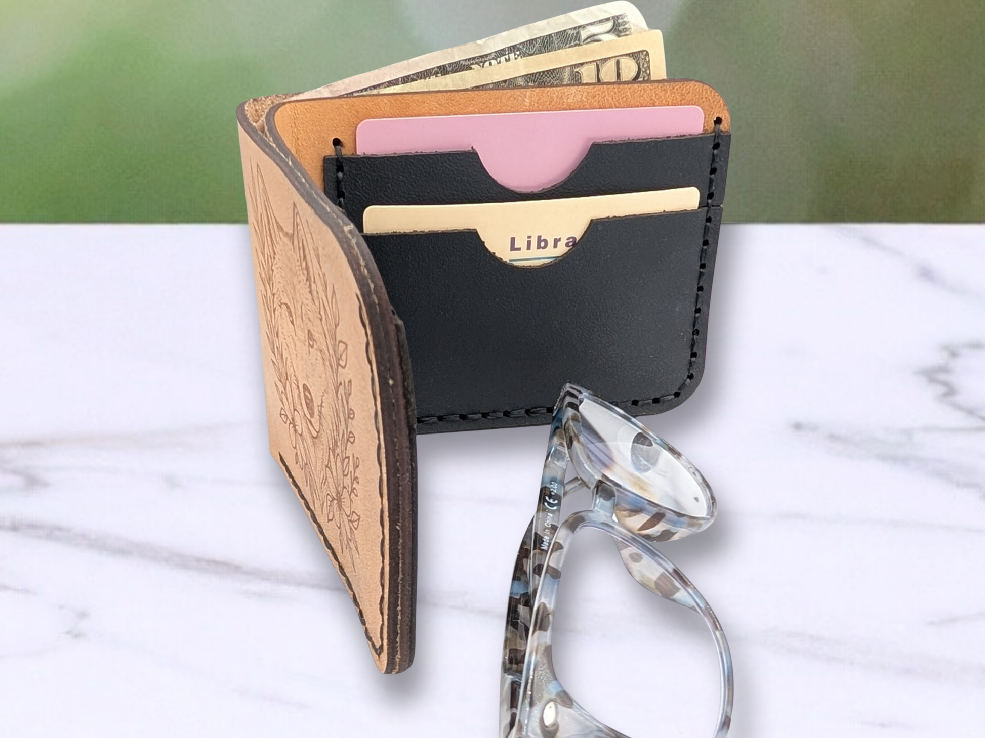 Bi Fold Veg Tan Wallet with wolf head laser engraved on the hand sewn wallet. Here the wallet is standing on the edge and opened so that you can see cash poking up out of the cash compartment and you can see two of the black leather card compartments on the back inside of the wallet with cards in them. The wallet has black stitching and the edges of the leather are dark brown. Glasses are in the photo to help with sizing. 