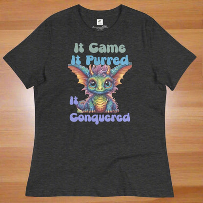 dark grey heather Women's Dragon T-Shirt flat on wooden counter. Green Iridescent baby kawaii dragon with large ears lined in orange. "It Came, It Purred, It Conquered" In Green light blue, and lavender.