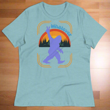 Heather Blue Lagoon Sasquatch Women's T-Shirt laying flat on a wooden surface. Big Foot in periwinkle against a rainbow and mountains showing a peace sign with text Whatever. Bigfoot Yeti Women's Tee