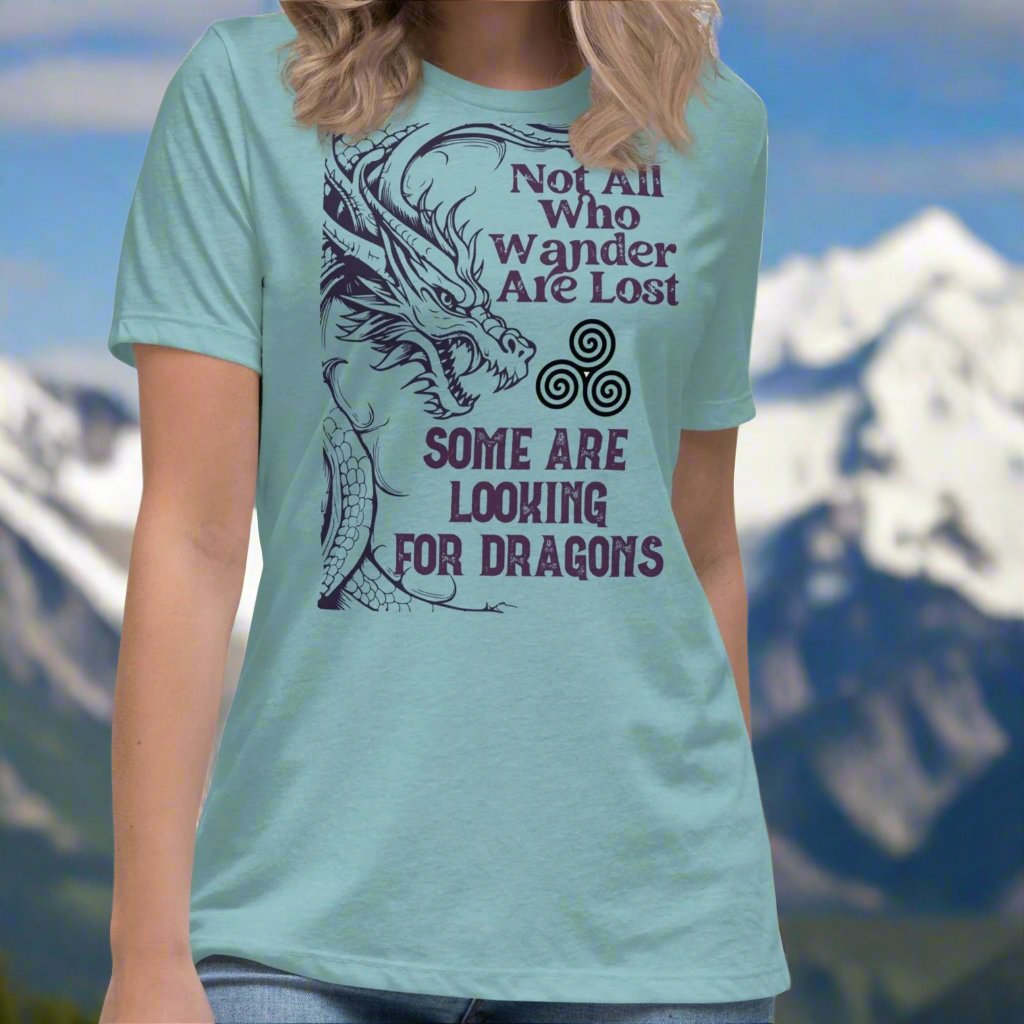 Heather Blue Lagoon, Front View, Women's T-Shirt. Right side is bordered a dragon in a grey purple. Looking like smoke coming from the dragons nostrils is the Celtic symbol triskele or the triple circle. Above this it says Not All Who Wander Are Lost. Below this it says Some Are Looking for Dragons.