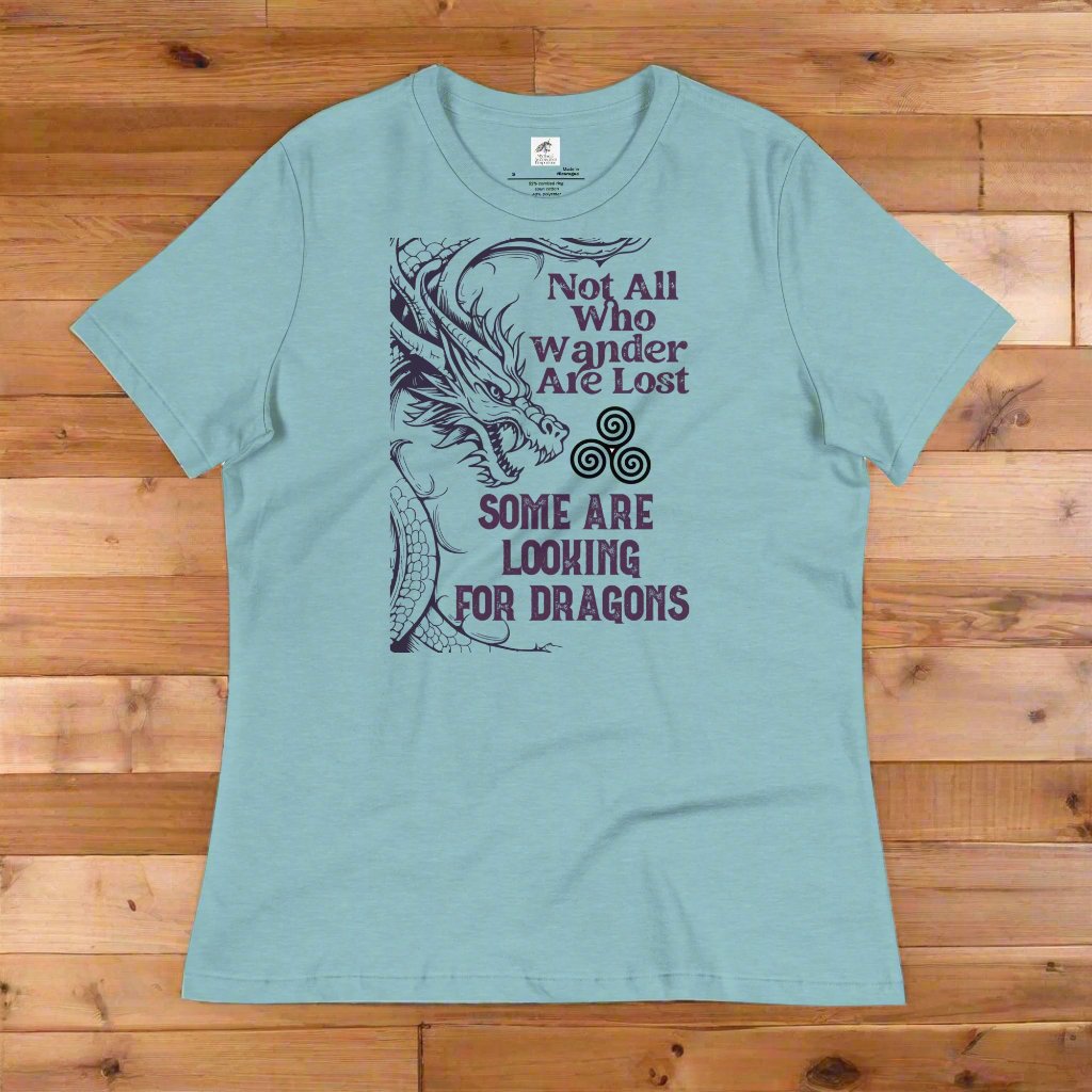 Heather Blue Lagoon, Front View on counter, Women's T-Shirt. Right side is bordered a dragon in a grey purple. Looking like smoke coming from the dragons nostrils is the Celtic symbol triskele or the triple circle. Above this it says Not All Who Wander Are Lost. Below this it says Some Are Looking for Dragons.