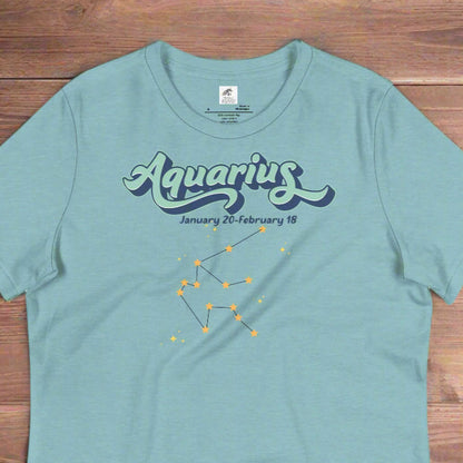 Flat lay heather blue lagoon astrology Aquarius women's T-shirt on a wooden counter. At the top in light blue is the word Aquarius outlined in deeper blue. Below this it says January 20- February 18. Under this is the Aquarius constellation with orange yellow stars connected by a black line. Astrology Zodiac Aquarius Woman's Tee