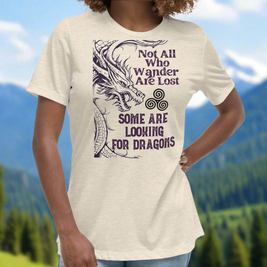 Heather Prism Natural, Front View, Women's T-Shirt. Right side is bordered a dragon in a grey purple. Looking like smoke coming from the dragons nostrils is the Celtic symbol triskele or the triple circle. Above this it says Not All Who Wander Are Lost. Below this it says Some Are Looking for Dragons.
