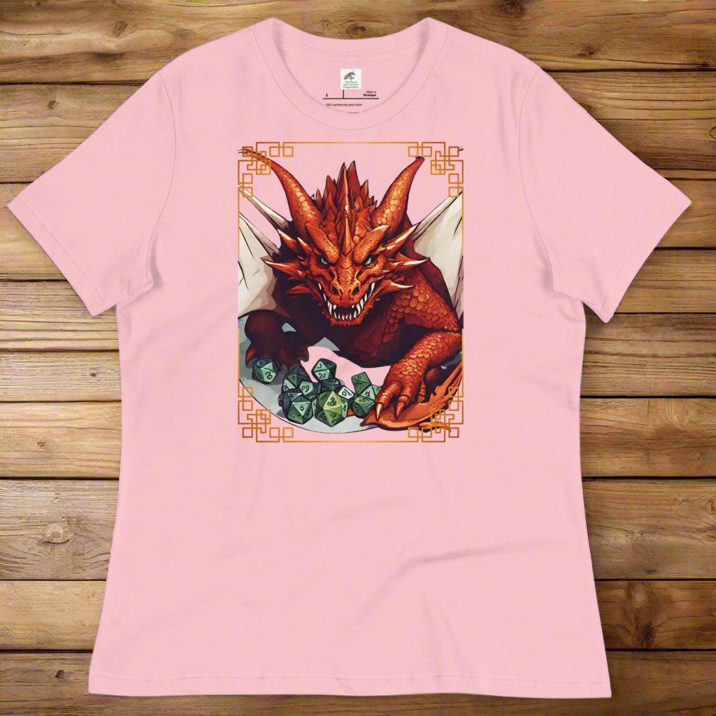 pink Front View Women's  T-Shirt laying flat on wood surface. DND players gift for adventurers. Red Dragon, wings spread, stalking over a pile of D20 and other dice. RPG and Dungeons Masters